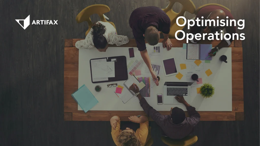 Two people sit at a desk reviewing a laptop screen that displays a colourful Artifax scheduling calendar. One person gestures toward the laptop while the other points with a green pencil. The text “How To Effectively Use Calendars” appears on the right side of the image.