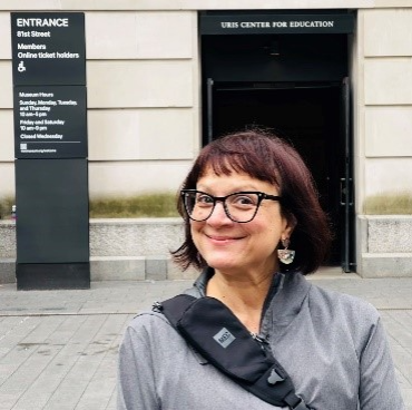 Woman smiling outside the entrance to a museum, wearing glasses and a shoulder bag.