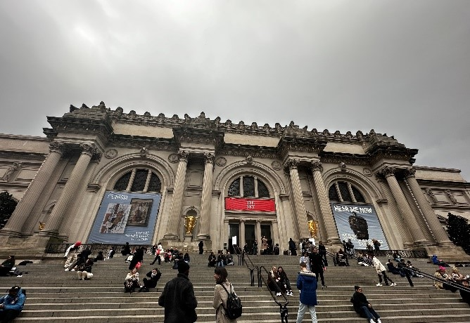 People sitting and walking on the steps of a large classical museum building with banners on the façade.