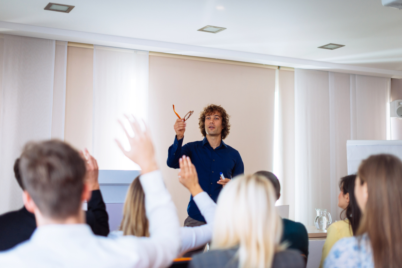 Presenter holding glasses while audience members raise their hands in a workshop setting.
