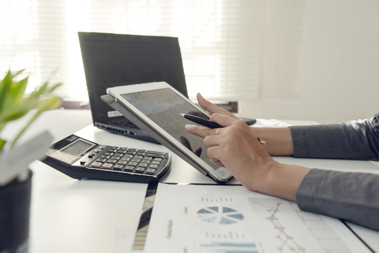 Hands using a tablet and calculator beside a laptop and printed financial charts on a desk