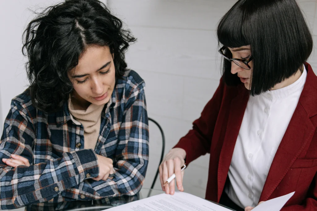 Two women reviewing a printed document at a table—one in a red blazer explaining a section while the other listens attentively, suggesting a one-on-one consultation or advisory session.