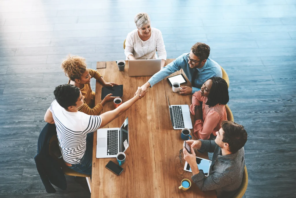 A diverse group of six colleagues sitting around a large wooden table, collaborating and shaking hands in a modern office setting.