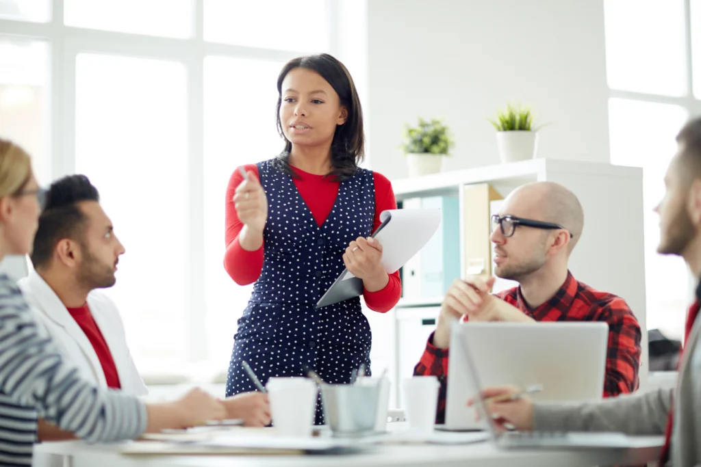 A confident woman leads a team discussion in a bright office space, holding a clipboard and gesturing while speaking to colleagues seated around a table.