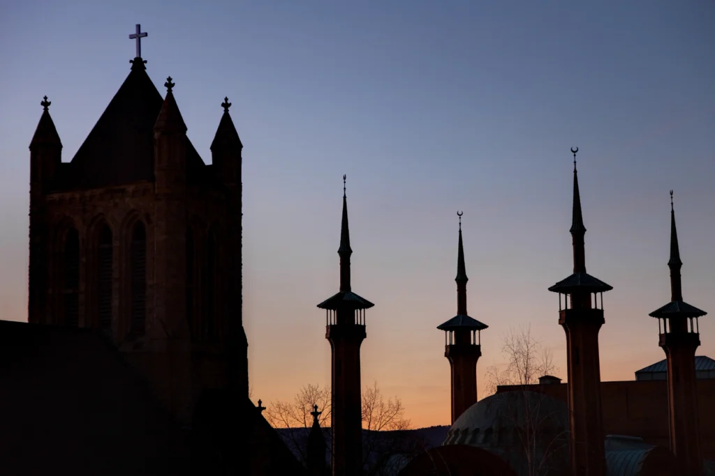 Silhouette of a tall tower with a cross and several slender spires with crescent decorations, set against a sunset sky.