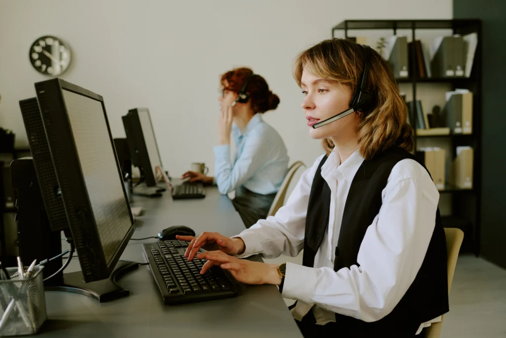 Two women working in a call center, wearing headsets and typing on desktop computers—depicting customer service, tech support, or helpdesk operations.