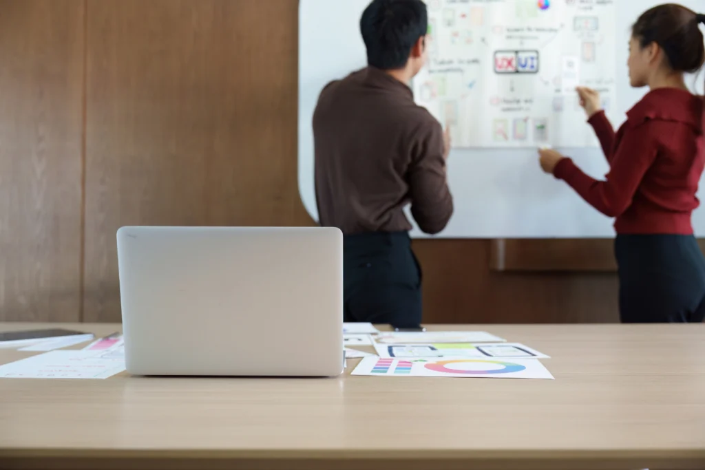 Two team members brainstorm at a whiteboard filled with UX/UI sketches and planning diagrams, with a laptop and charts spread out on a desk in the foreground.