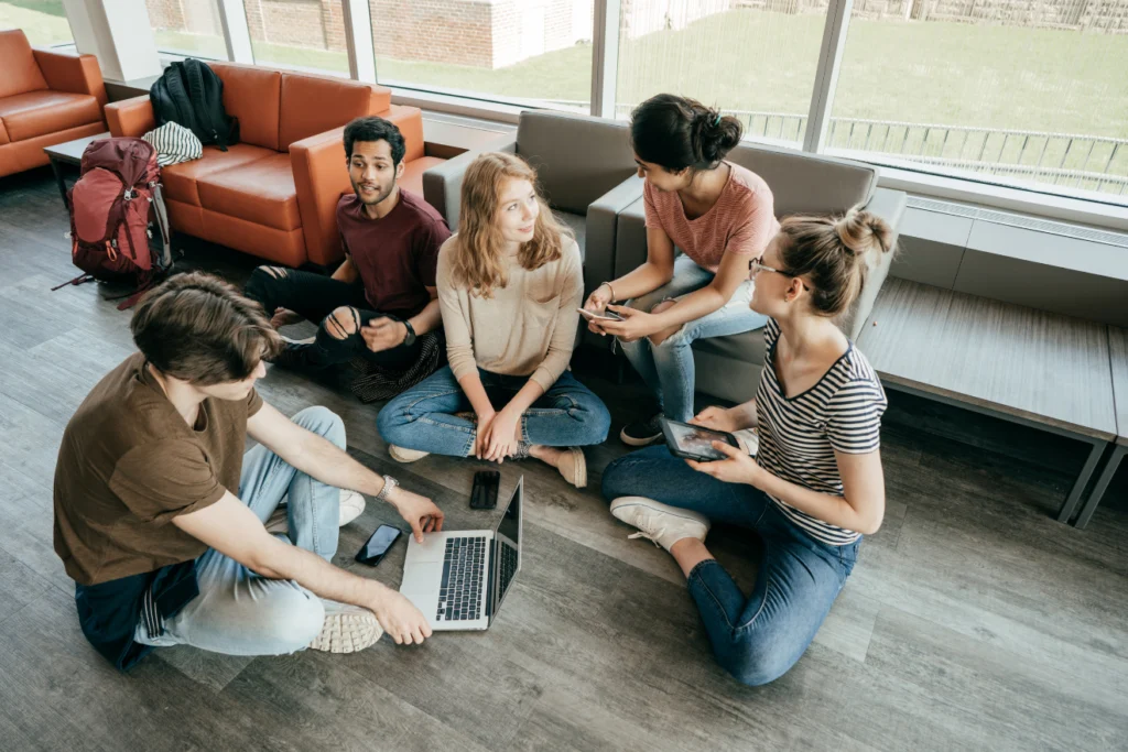A group of young adults casually seated on the floor in a lounge area, chatting and using digital devices.