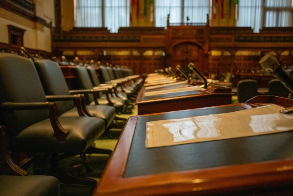 Empty council chamber with rows of desks equipped with microphones and papers.