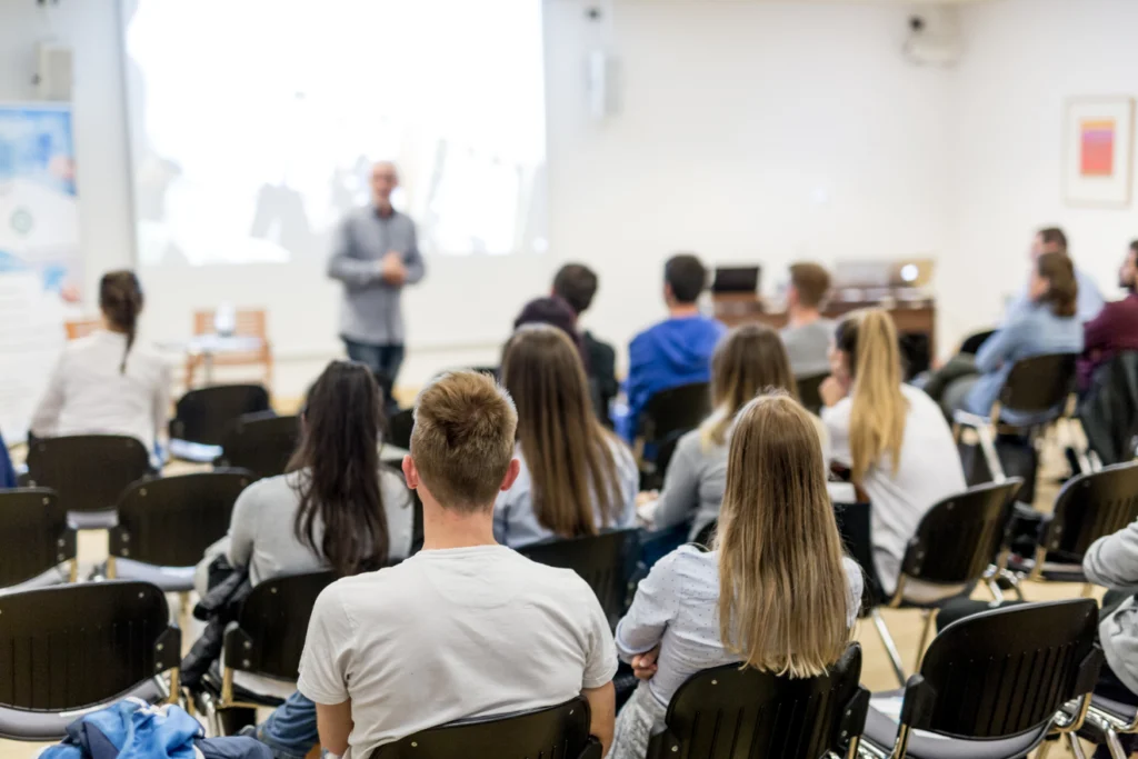 Audience seated in a classroom watching a speaker present at the front of the room.