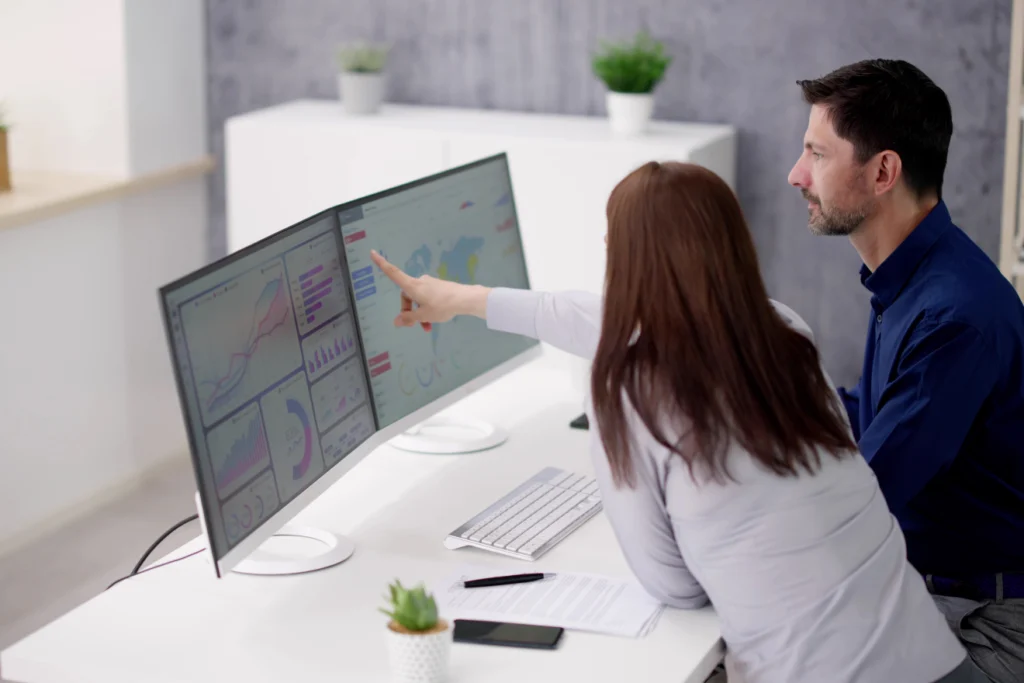 A woman points at a large dual-monitor setup displaying colorful data dashboards and maps, while a male colleague looks on—suggesting collaboration on performance or analytics insights.