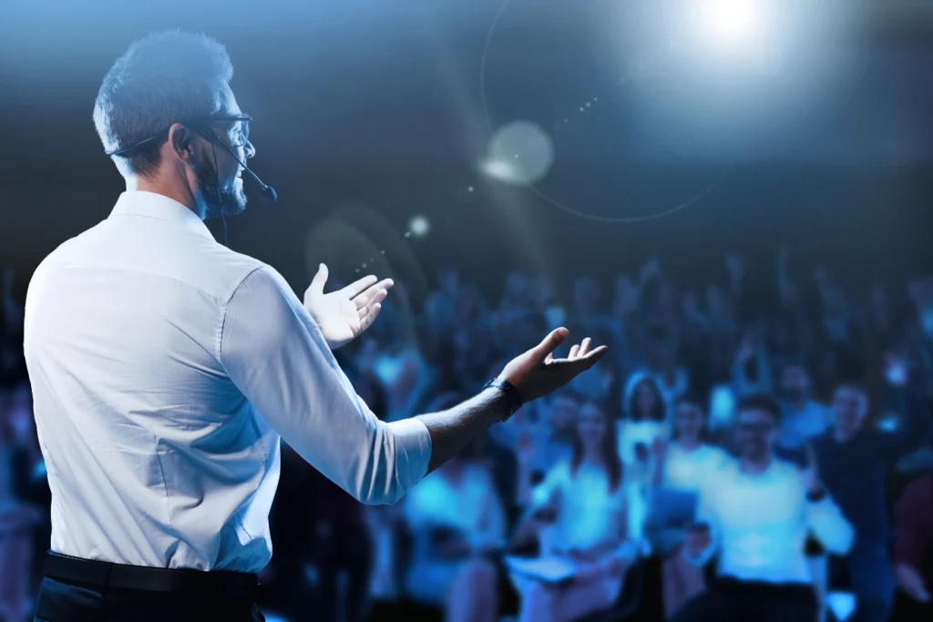 Speaker with headset microphone addressing an excited audience in blue lighting.