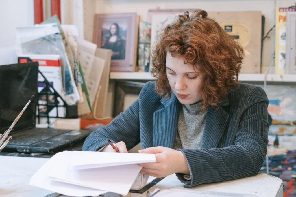 Artist reading and marking papers at a cluttered studio desk.