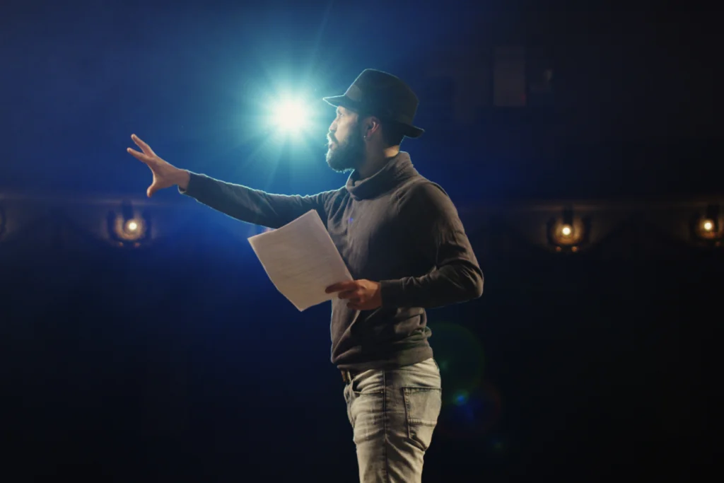 Man rehearsing on stage under a spotlight, holding a script.