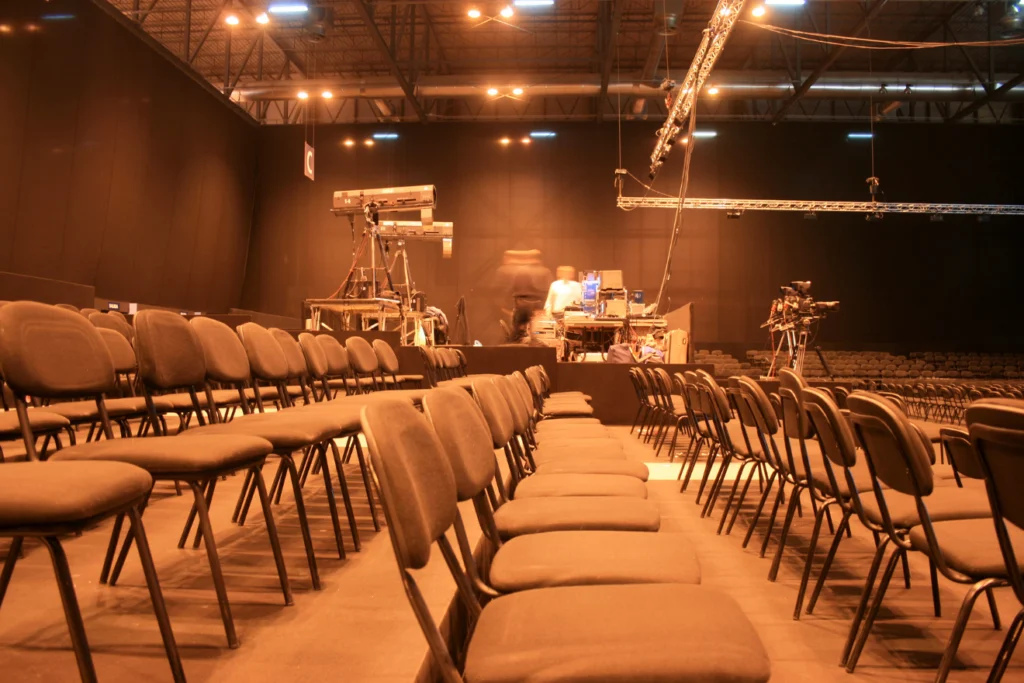 Rows of empty conference chairs with AV equipment and cameras behind.