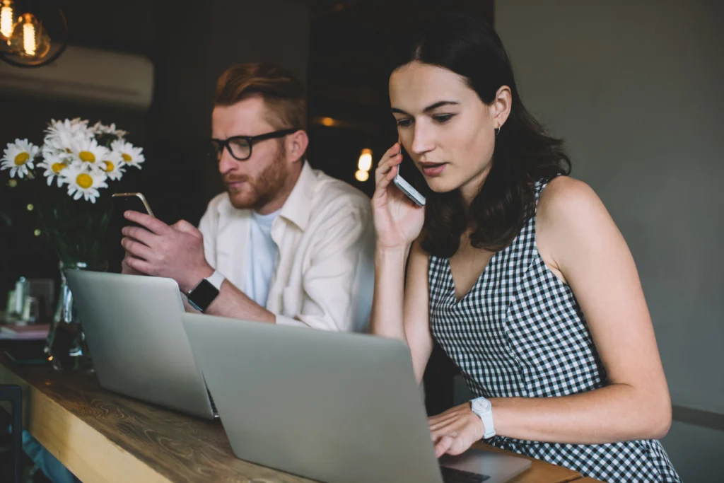 Two coworkers use laptops and smartphones while working at a shared table.