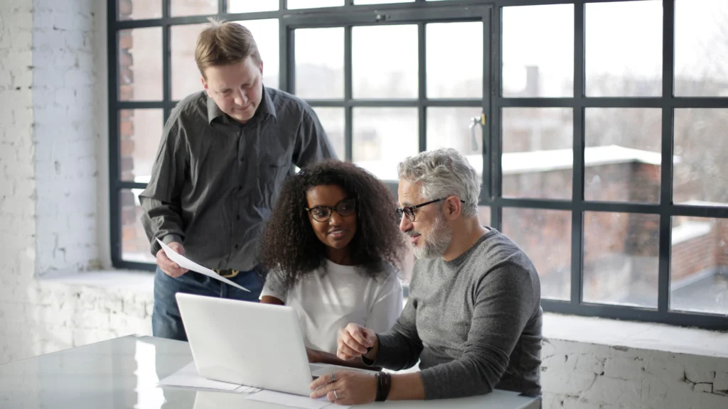 Three coworkers collaborating around a laptop in a modern office with large windows.