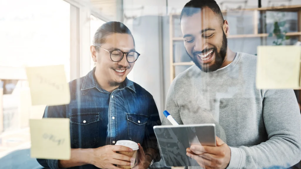 Two men brainstorming with sticky notes on a glass wall, smiling while looking at a tablet.