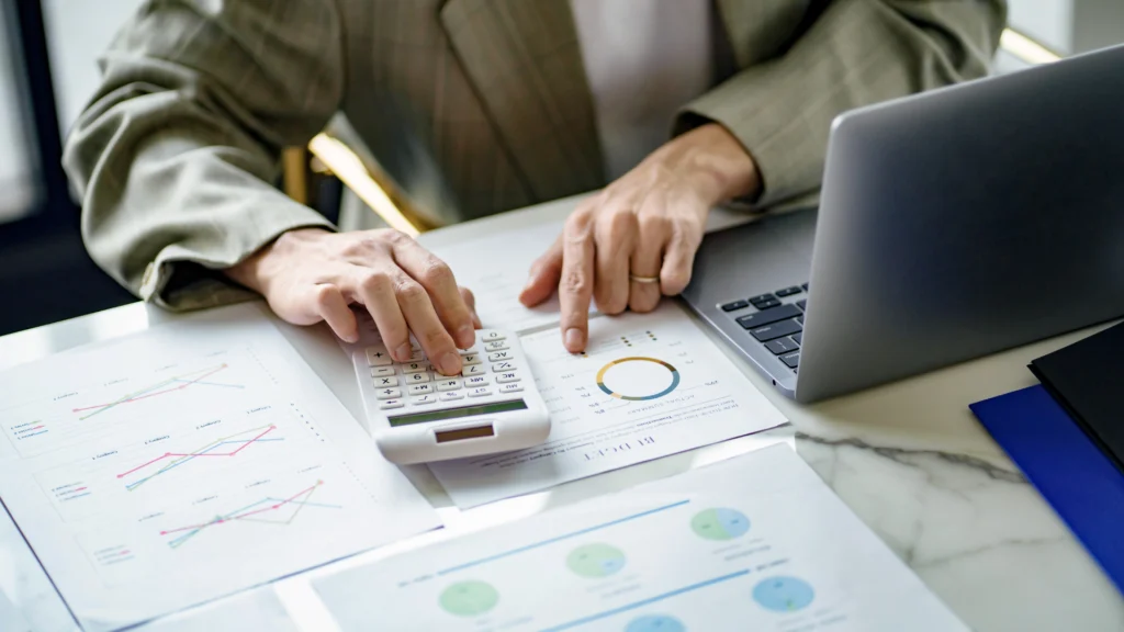 Person using a calculator and reviewing financial charts next to a laptop on a desk.