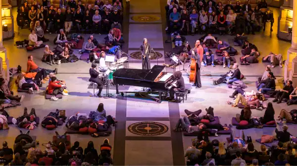 Musicians perform on a stage with a grand piano in a large hall while a crowd sits and lies on the floor around them, listening.