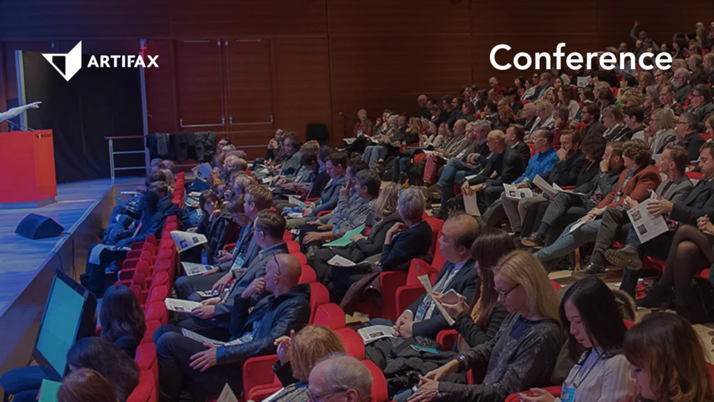 Audience seated in a modern auditorium attending a conference, with a speaker at a podium and the Artifax logo displayed in the top left corner.