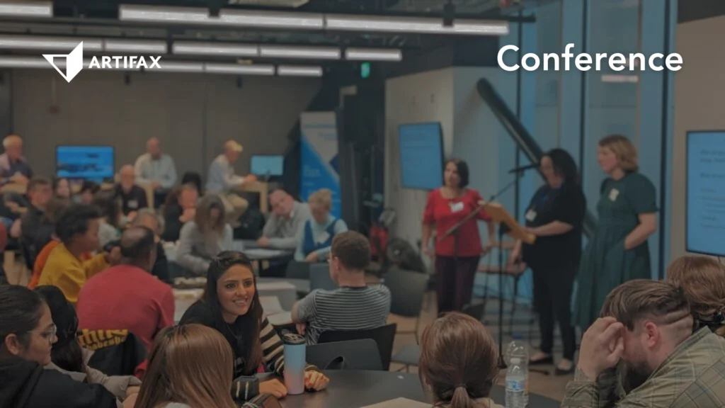 A roundtable-style conference with attendees engaged in discussion while presenters stand near a screen at the front of the room. Artifax logo and the word "Conference" are visible.
