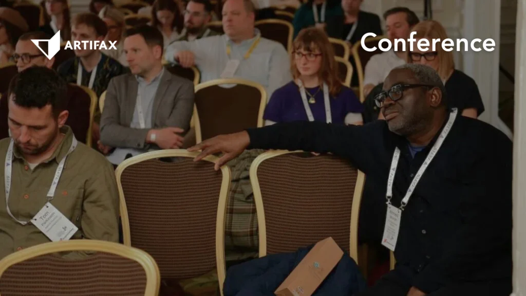 Seated attendees in a conference session, with one participant leaning across chairs mid-conversation. Artifax logo and the word "Conference" appear in the top corners.