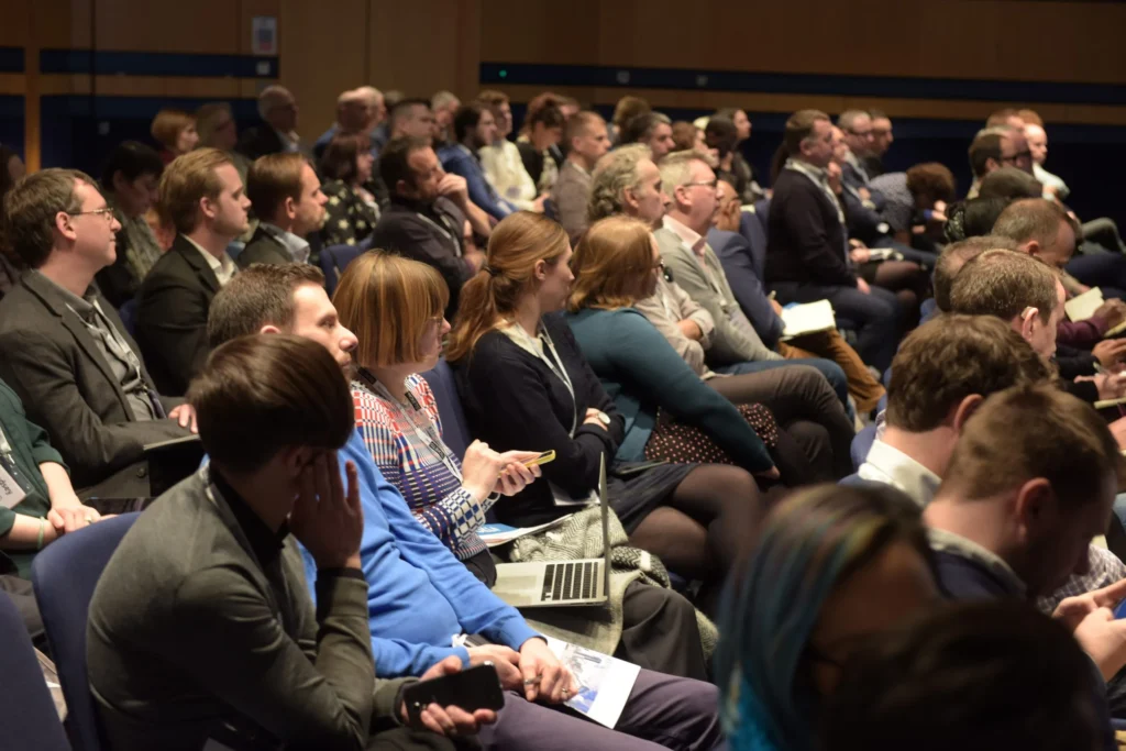 A full room of delegates attentively listening during a theatre-style conference presentation, with rows of seated participants facing the stage.