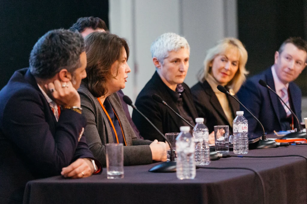 Panel of six professionals seated at a table with microphones and water bottles, speaking during a conference session.