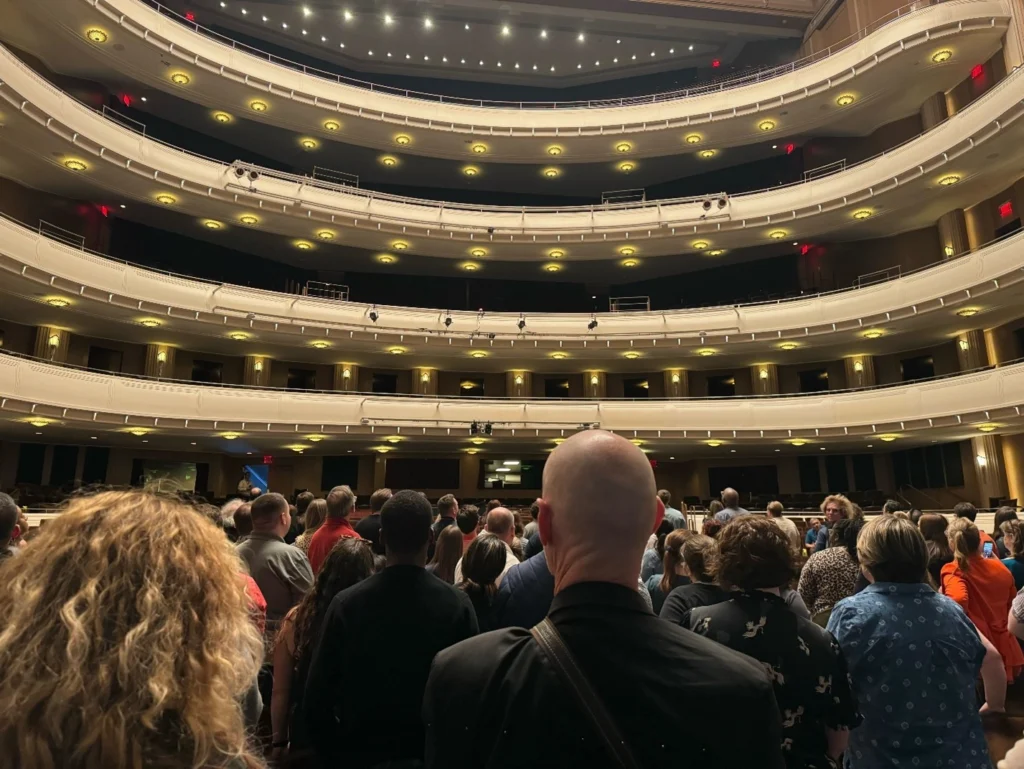 View from the stage looking out at a grand, multi-tiered performance hall filled with attendees standing and observing the architecture.