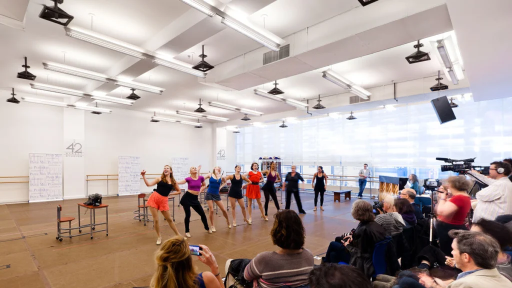 Performers rehearsing a musical number inside a bright studio at New 42 Studios while an audience watches.